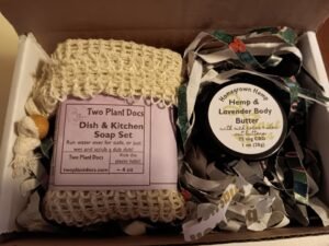 An open gift box containing a soap inside a sisal scrubbie/soap saver and a jar of body butter